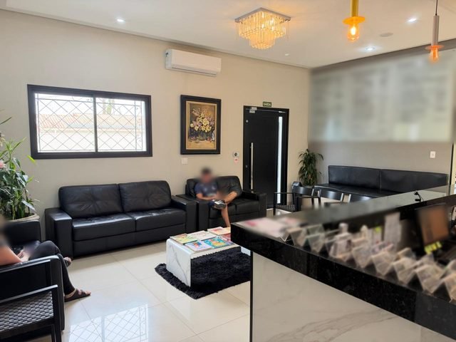 Sala de espera do prédio, com sofás de couro preto, cadeiras, janela com grade branca e balcão de recepção; rostos desfocados